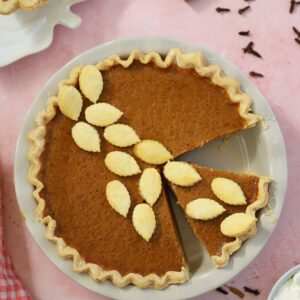 A gluten free pumpkin pie on a pink backdrop with cinnamon sticks and cloves surrounding it.