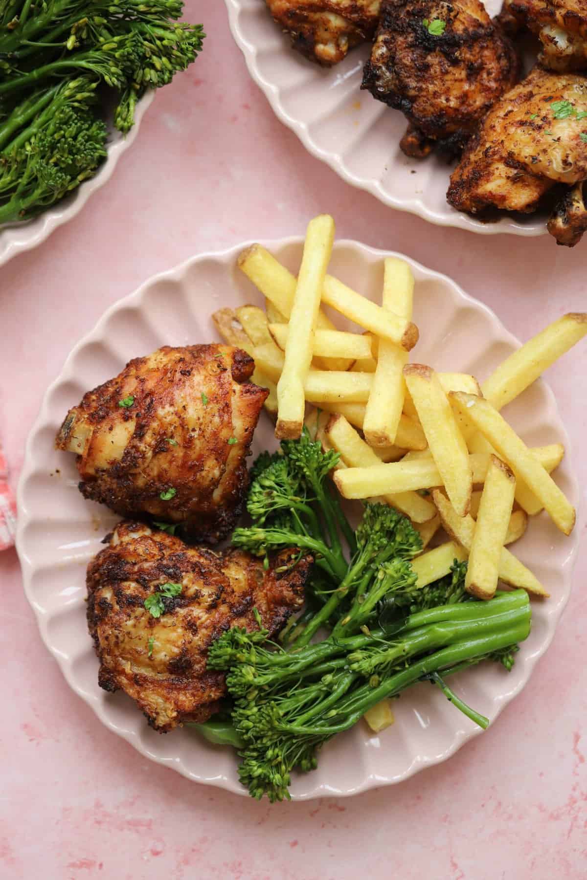 A plate of air fried chicken thighs with oven chips and tenderstem broccoli on a pink backdrop.