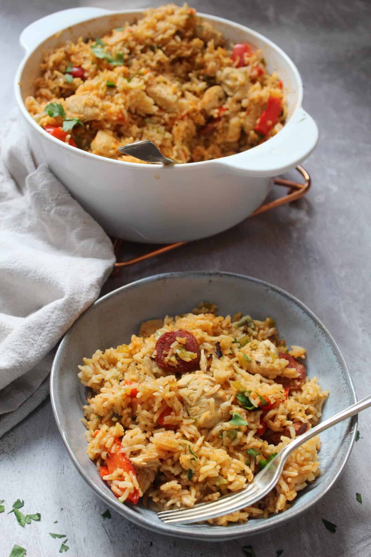 A bowl of jambalaya with chicken, chorizo and red peppers with a pan of jambalaya behind.