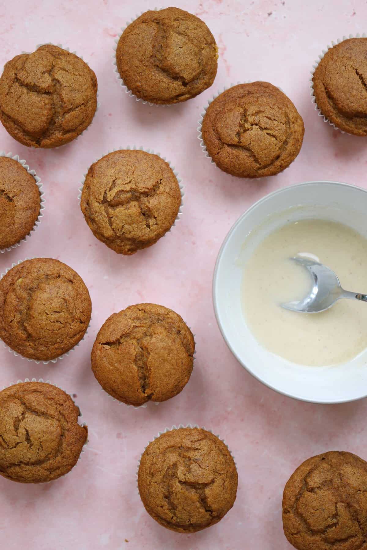 The pumpkin banana muffins on a pink worktop with a bowl of maple cinnamon glaze.