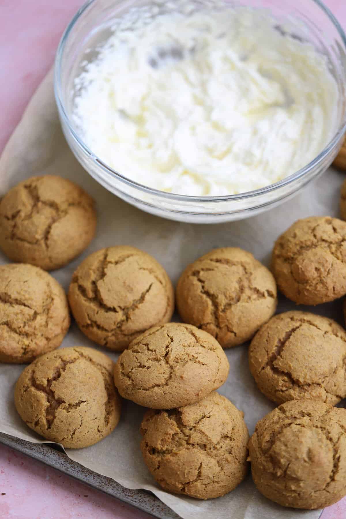 A baking tray with soft gluten free pumpkin cookies and a bowl of cream cheese frosting.