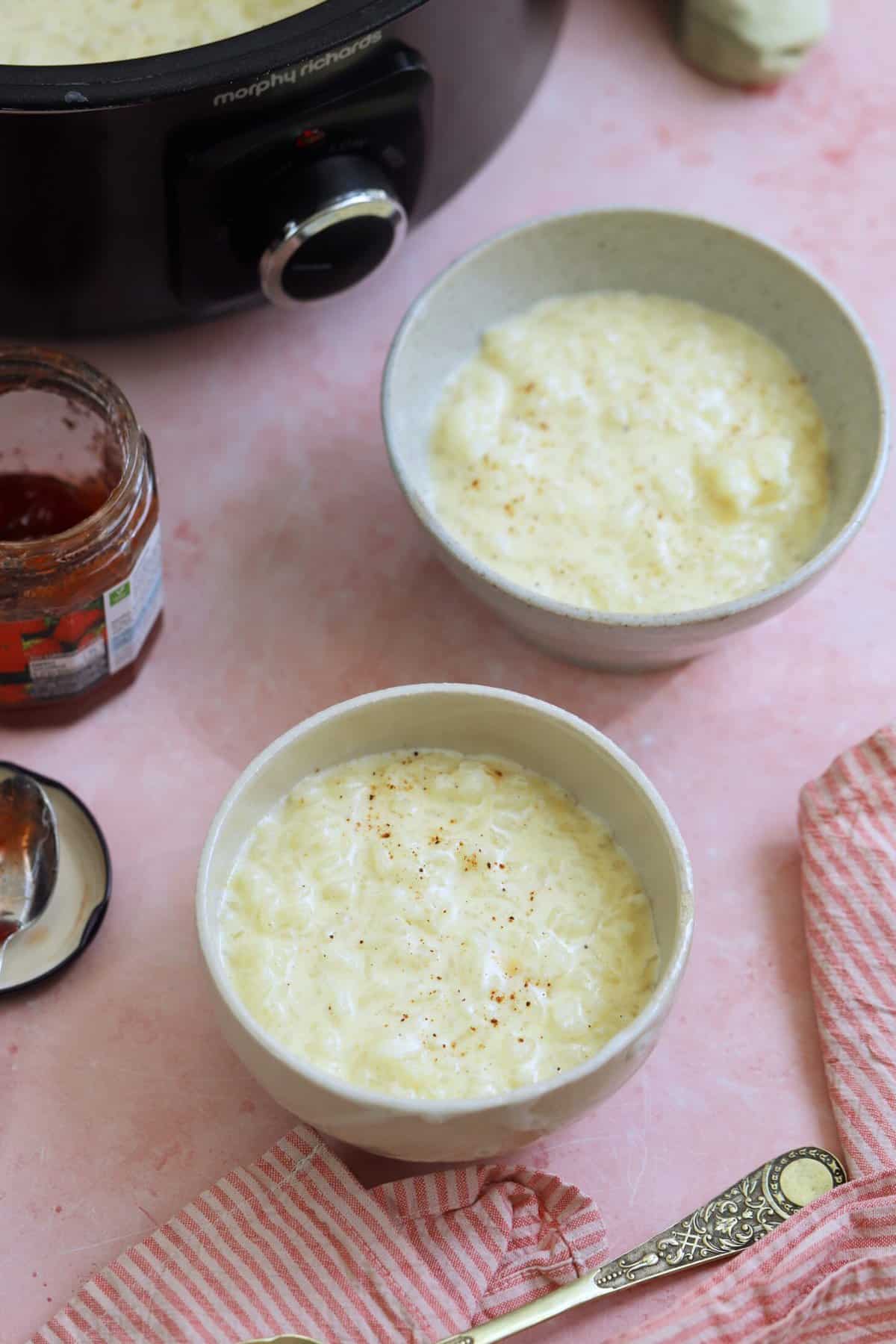 Two bowls of slow cooker rice pudding in front of a crock pot and an open jar of jam.